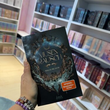 Buchcover von 'Musenrausch' von Malou Bichon in einer Hand gehalten, umgeben von Bücherregalen.