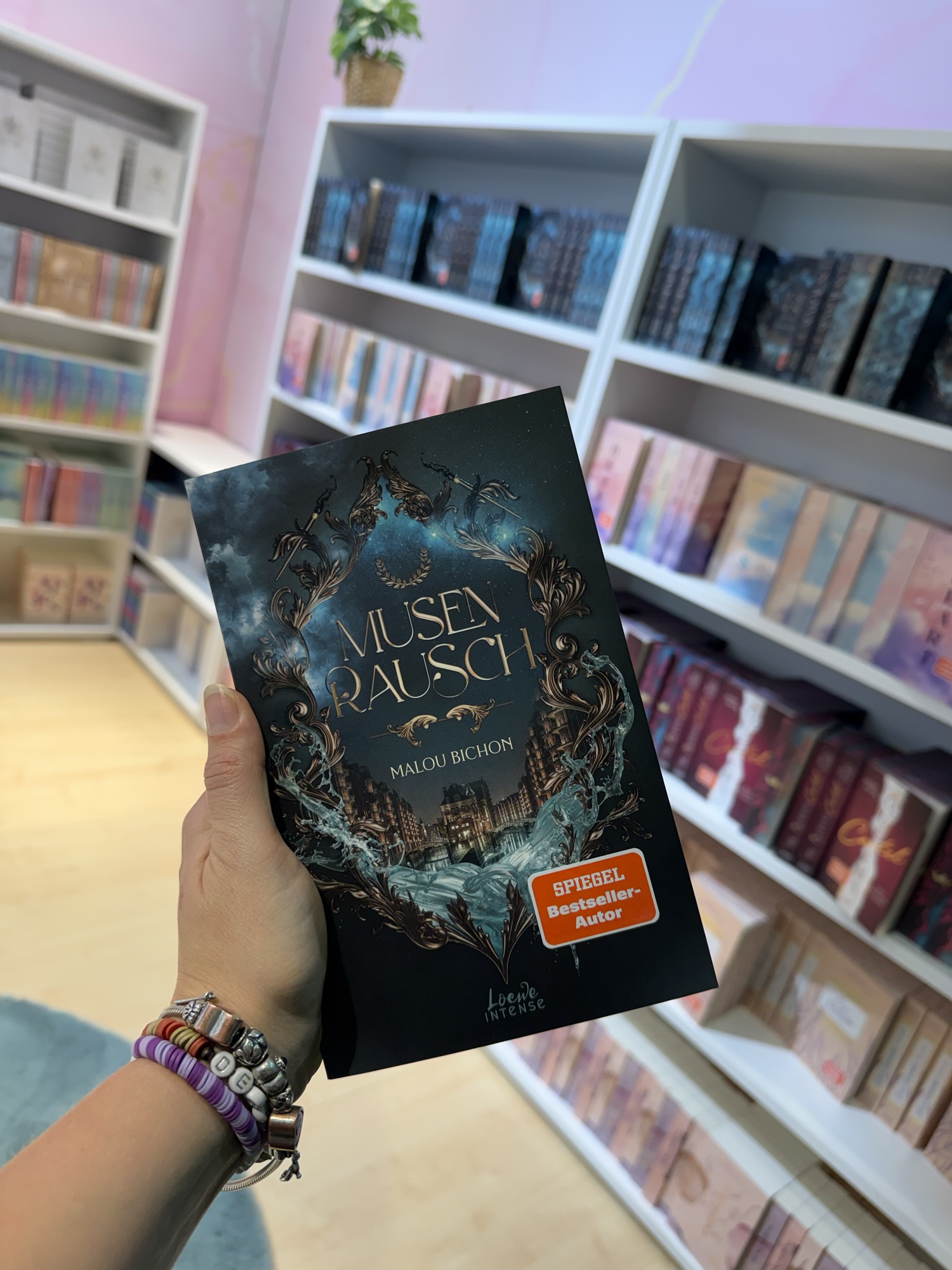 Buchcover von 'Musenrausch' von Malou Bichon in einer Hand gehalten, umgeben von Bücherregalen.