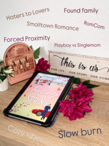 Auf einem Holztisch liegen ein Tablet mit „A Feeling of Spring“, eine Buchdeko und pinke Blüten, während Buchgenres wie RomCom und Slow Burn im Raum stehen.