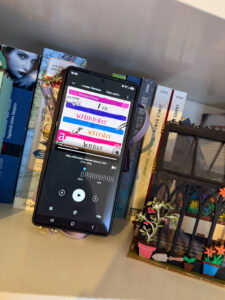Ein Smartphone zeigt die Audiobook-App mit „Mein schlimmster schönster Sommer“, umgeben von bunten Buchrücken.
