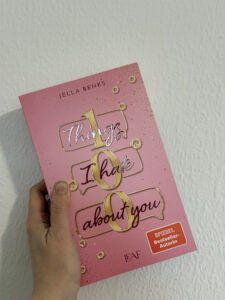 Eine Hand hält den rosafarbenen Roman „Things I Want to Say About You“ von Jella Benks mit goldener Schrift.