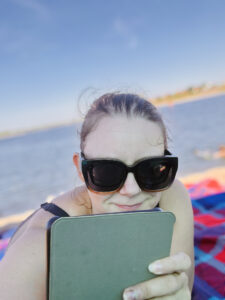 Eine Frau mit schwarzer Sonnenbrille liest am Strand konzentriert auf einem grünen Tablet, während hinter ihr das Meer glitzert.