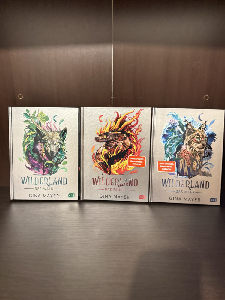 Drei B&auml;nde der Fantasy-Reihe Wilderland von Gina Mayer stehen nebeneinander und zeigen farbenfrohe, tierische Illustrationen.