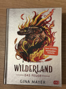 Drei Bände der Fantasy-Reihe Wilderland von Gina Mayer stehen nebeneinander und zeigen farbenprächtige Tierwesen auf den Buchcovern.