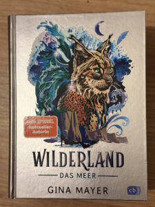 Das Buchcover von „Wilderland – Das Meer“ zeigt einen detailreich illustrierten Luchs mit magischen Wellen, Mond und Feder.